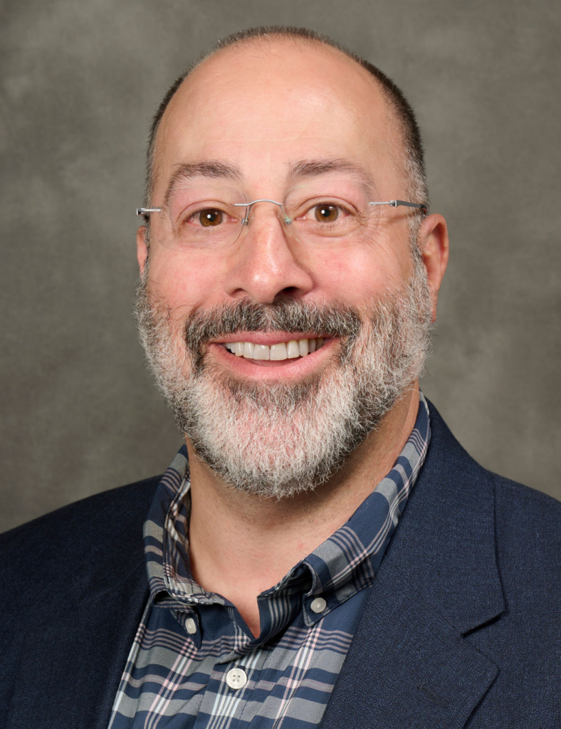 Smiling man with glasses and a gray beard in a blue plaid shirt and navy blazer, neutral backdrop behind him.